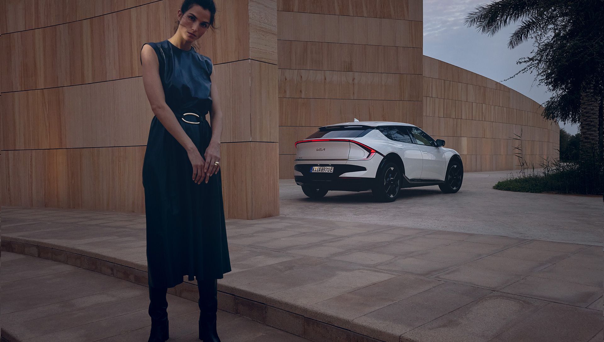 A woman stands beside a Kia EV6 in front of a modern building, showcasing the car's sleek design and urban setting.